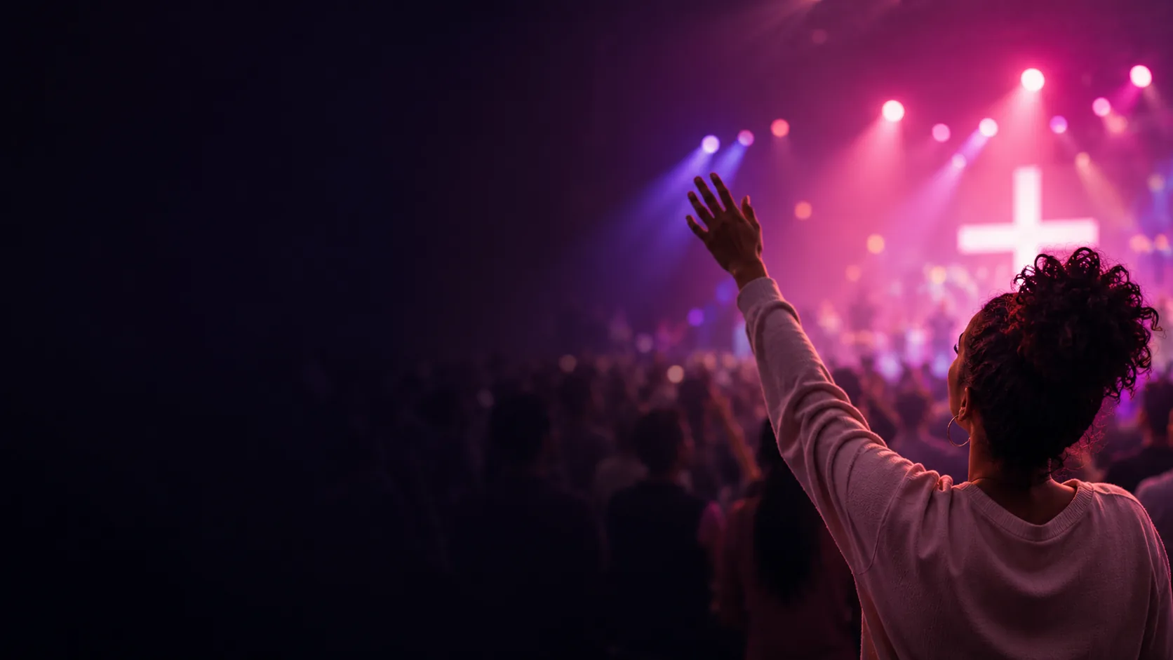 A woman raises her hand in worship facing a stage glowing with pink light and a cross